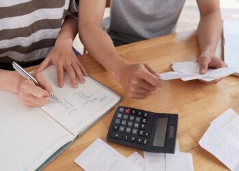 Close-up shot of unrecognizable family gathered together at wooden table and keeping records of expenses, woman taking notes in notepad
