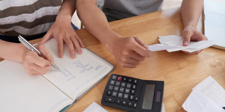 Close-up shot of unrecognizable family gathered together at wooden table and keeping records of expenses, woman taking notes in notepad