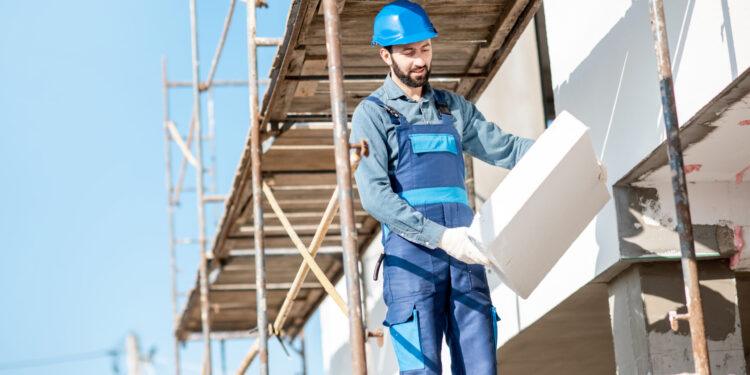 Builder warming a building facade with foam panels standing on the scaffoldings on the construction site