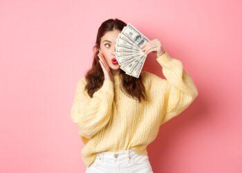 Coquettish young lady pucker lips, making silly face, covering half of it with money dollars, standing over pink background. Copy space