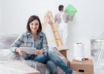 Happy woman relaxing and connecting with her tablet while a painter is painting the room, home renovation concept