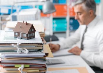 Real estate agent working in the office and piles of paperwork, model house on the foreground and mortgage loan documentation
