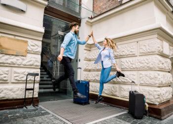 Young cheerful couple with suitcases jumping and giving five, having fun, before arriving to the hotel. Couple having fun outdoors, near the old city building.
